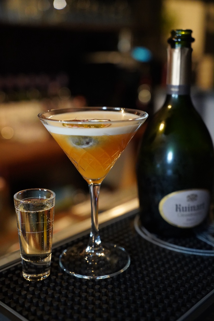 Cocktail mit Schaumhaube neben einem Shot und einer Flasche Ruinart